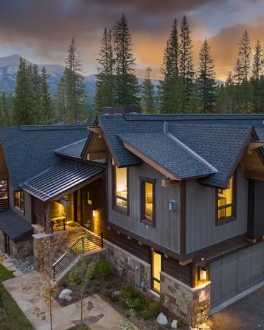 Modern mountain home with dark metal roof and stone foundation surrounded by tall evergreen trees and mountains at sunset