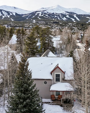 Snow-covered house with ski slopes and mountains visible in the background, surrounded by evergreen and aspen trees