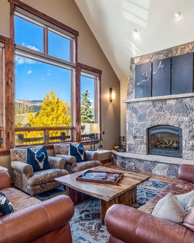 Spacious living room with burgundy leather seating, stone fireplace, and large windows overlooking mountains and yellow fall foliage