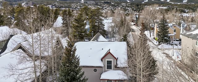 Snow-covered house with red roof peak surrounded by bare trees, ski slopes visible on mountains in background